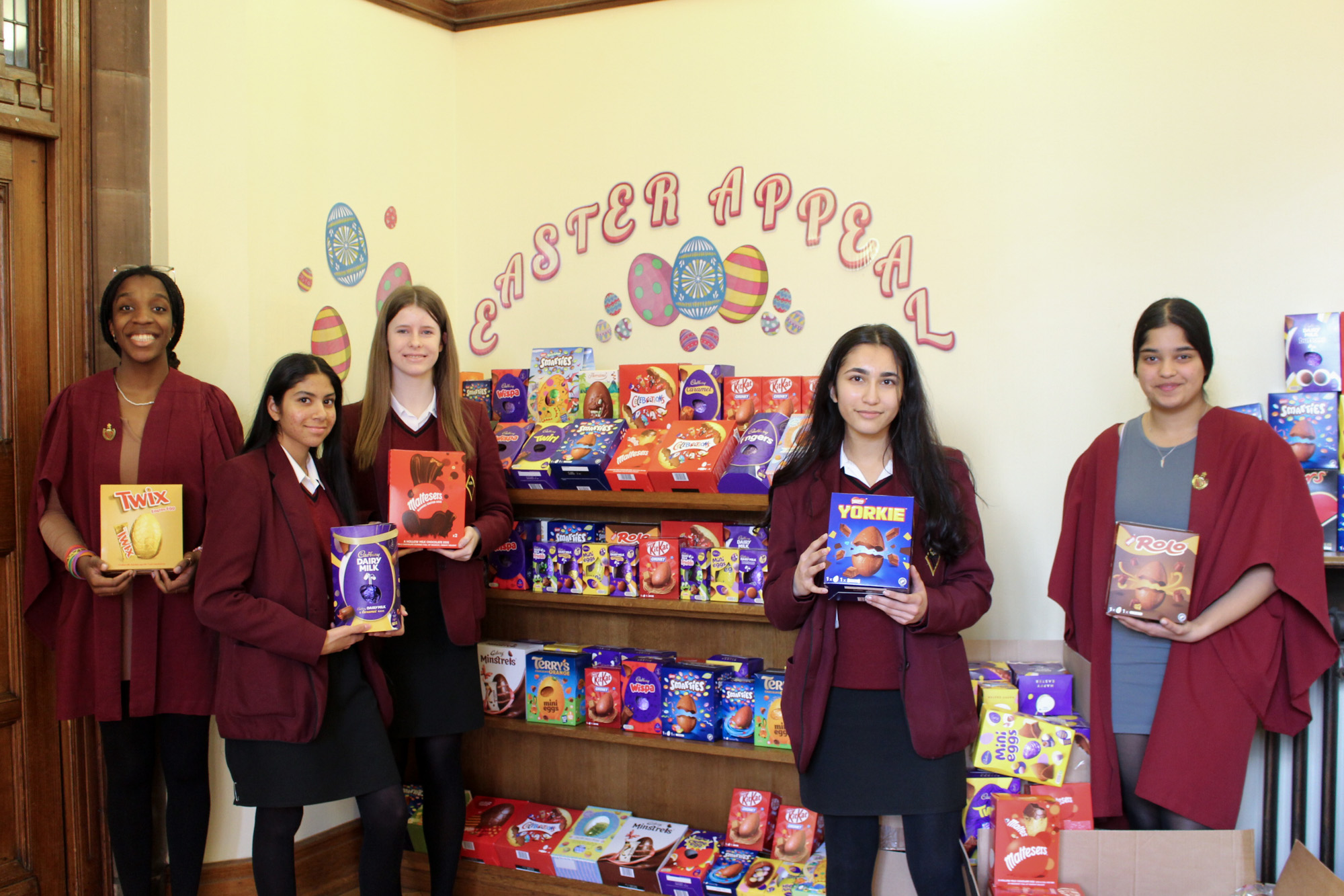 5 Bolton School Girls' Division pupils standing in front of a large and full 'Easter Appeal' display of Easter eggs, featuring a colourful array of boxed chocolate eggs.