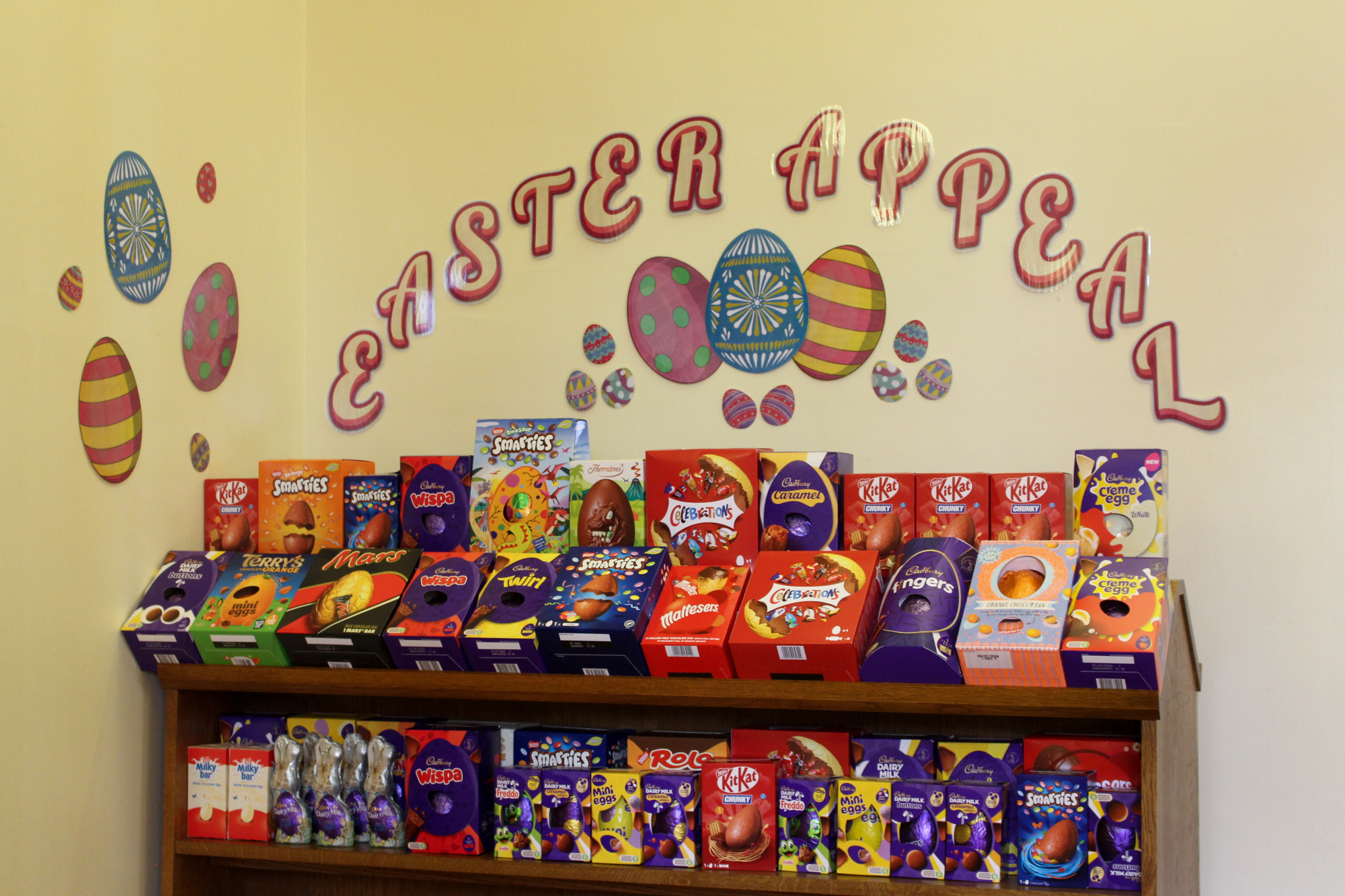A set of shelves full to bursting with varied and colourful boxed Easter Eggs, above which the words 'Easter Appeal' have been stuck to the wall in an arch, with images of colourful eggs stuck under the words and to the side.
