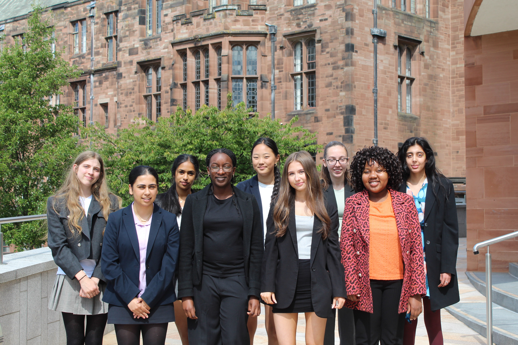 A diverse group of 9 smartly dressed Sixth Form girls standing outside in the sunshine with the red sandstone buildings of Bolton School Girls' Division and the Riley Sixth Form Centre in the background.