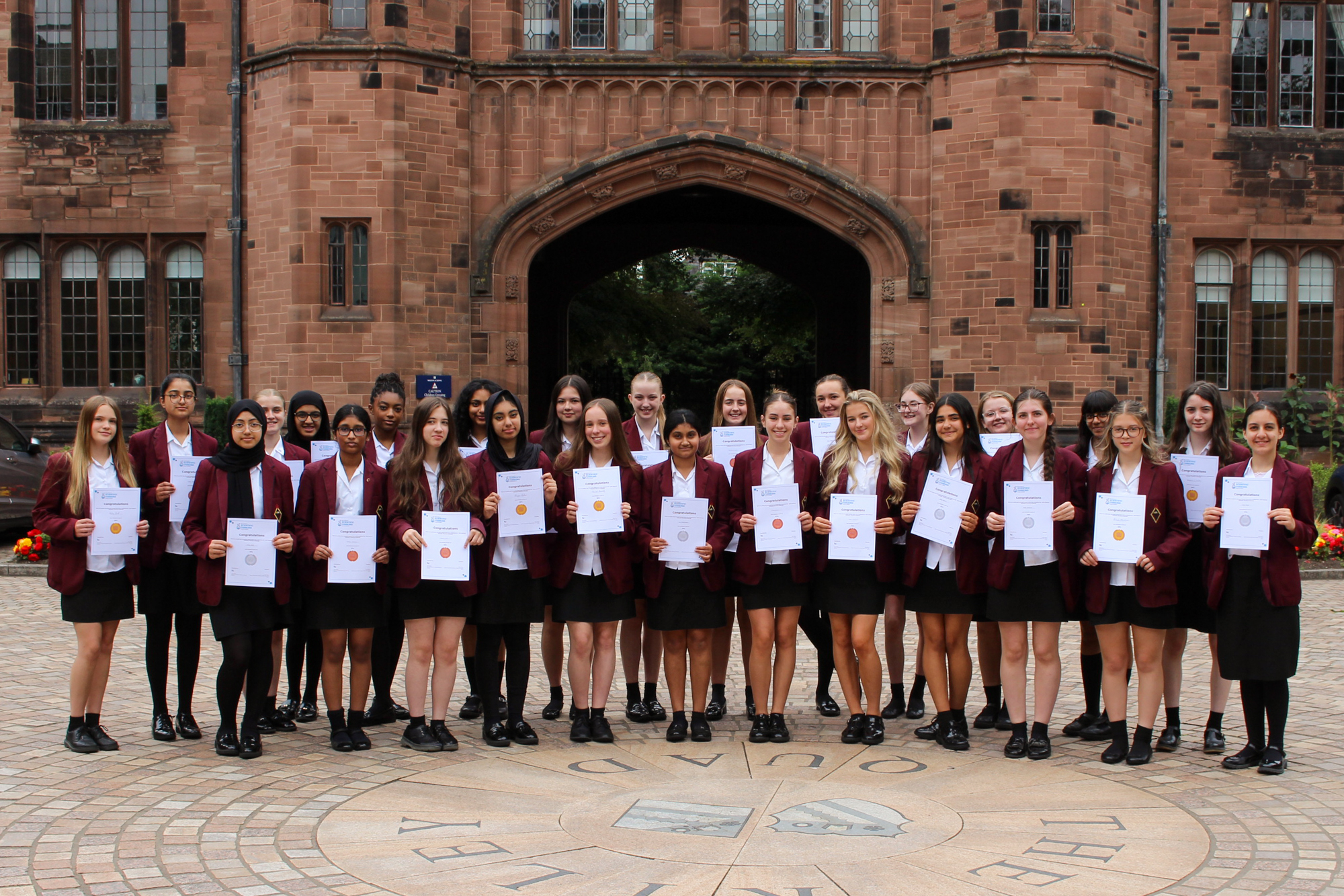 A large group of Year 10 girls in school uniforms with maroon blazers and proudly holding certificates while standing in front of Bolton School's iconic red sandstone archway