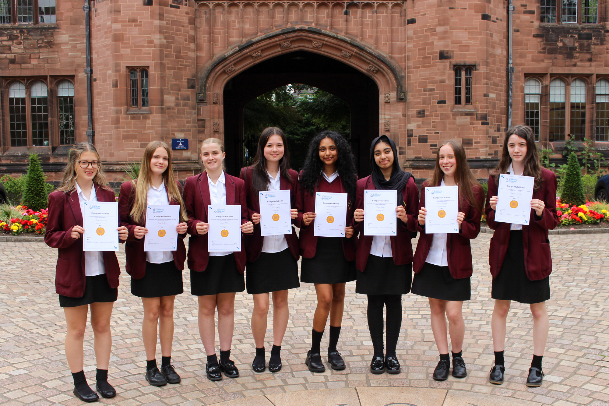 A group of 8 smiling Year 10 girls holding up "gold" certificates and wearing school uniforms with maroon blazers, standing in front of the iconic red sandstone main arch of Bolton School