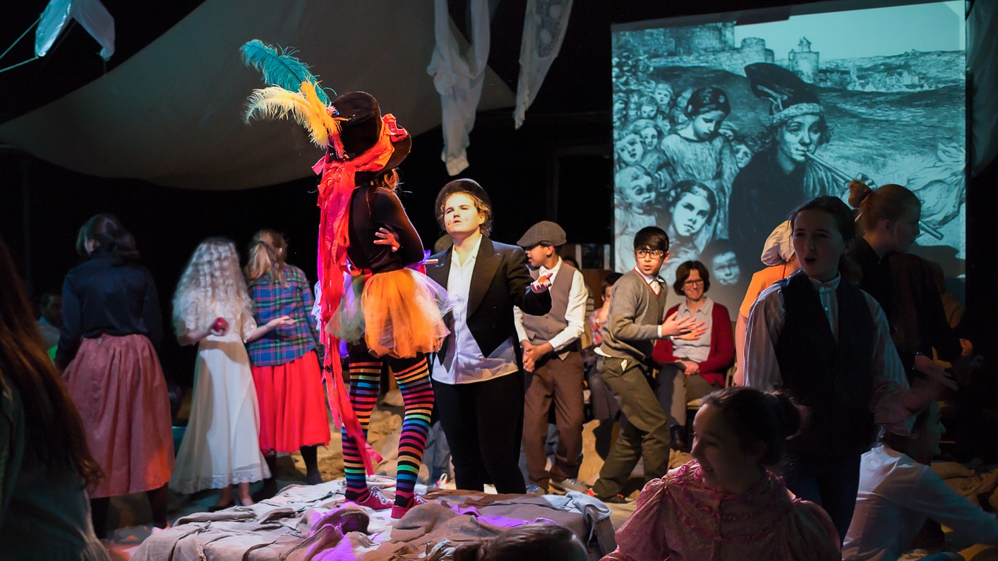 Rats Tales: A colourfully dressed figure in a top hat with feathers stands with arms folded at centre stage on a platform, with the rest of the cast fanned out around them