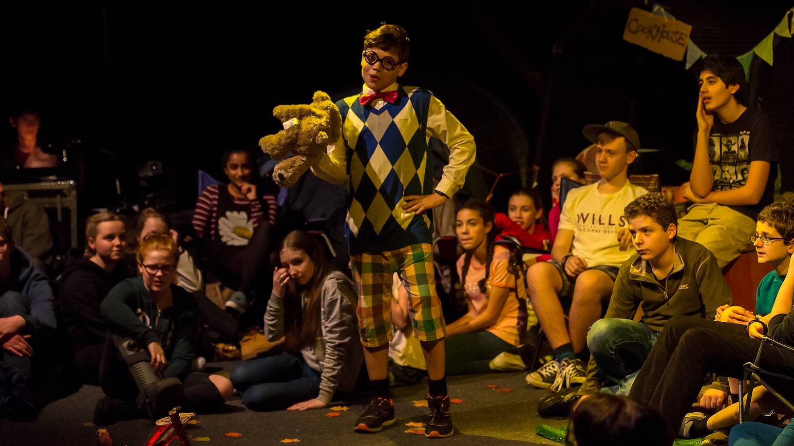 Wonder: a boy in mismatched patterns and prominent round glasses stands with a teddy under one arm and his hand on his hip, addressing the other cast members who are gathered around as if by a campfire to listen