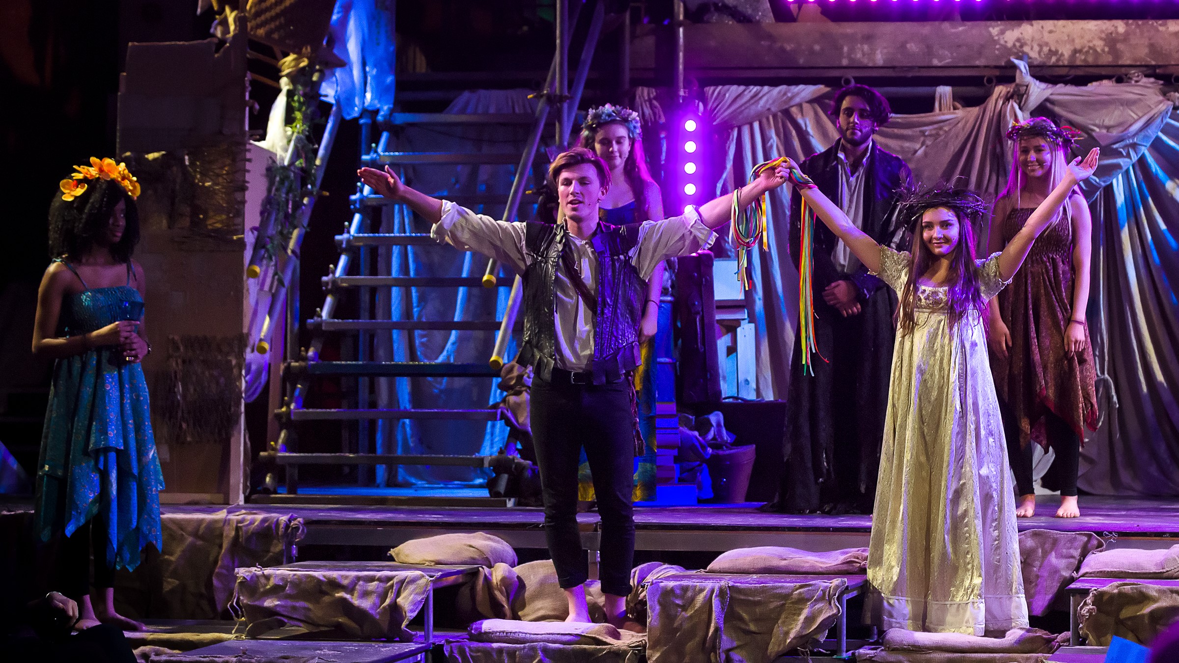 The Tempest: Ferdinand and Miranda stand with arms raised, the hand held between them bound by colourful thread as part of a wedding ceremony; they are surrounded by colourful "spirits" wearing crowns of flowers and leaves