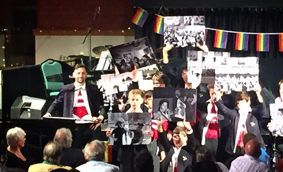 A group of students holding photos and placards, surrounded by rainbow flags, performing in the Arts Centre alongside Ralf Little