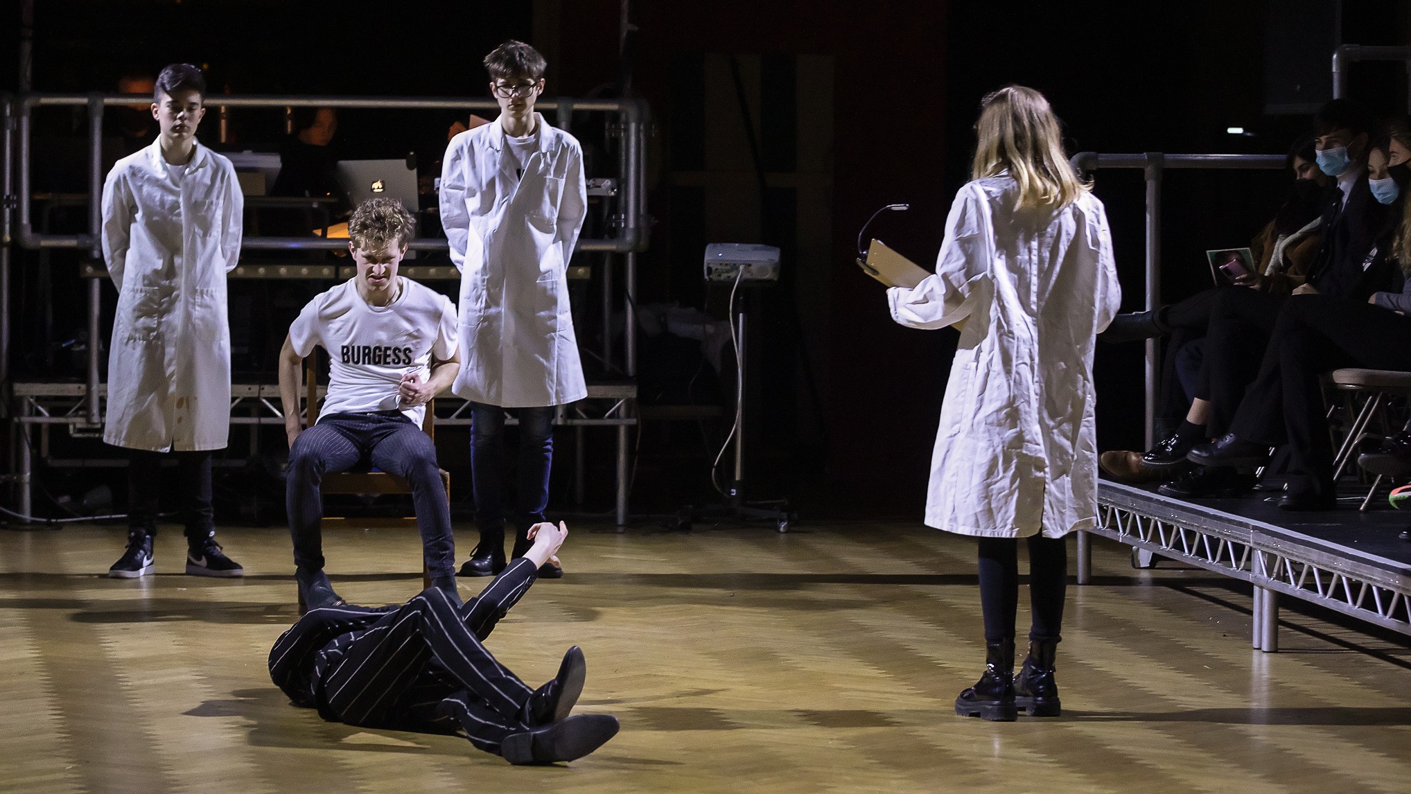 Clockwork: a character in a pinstripe suit lies on the floor at the feet of a characters in a white t-shirt that reads "Burgess" who looks disgusted; behind him are two boys in lab coats, and a girl in a lab coat stands by with a clipboard