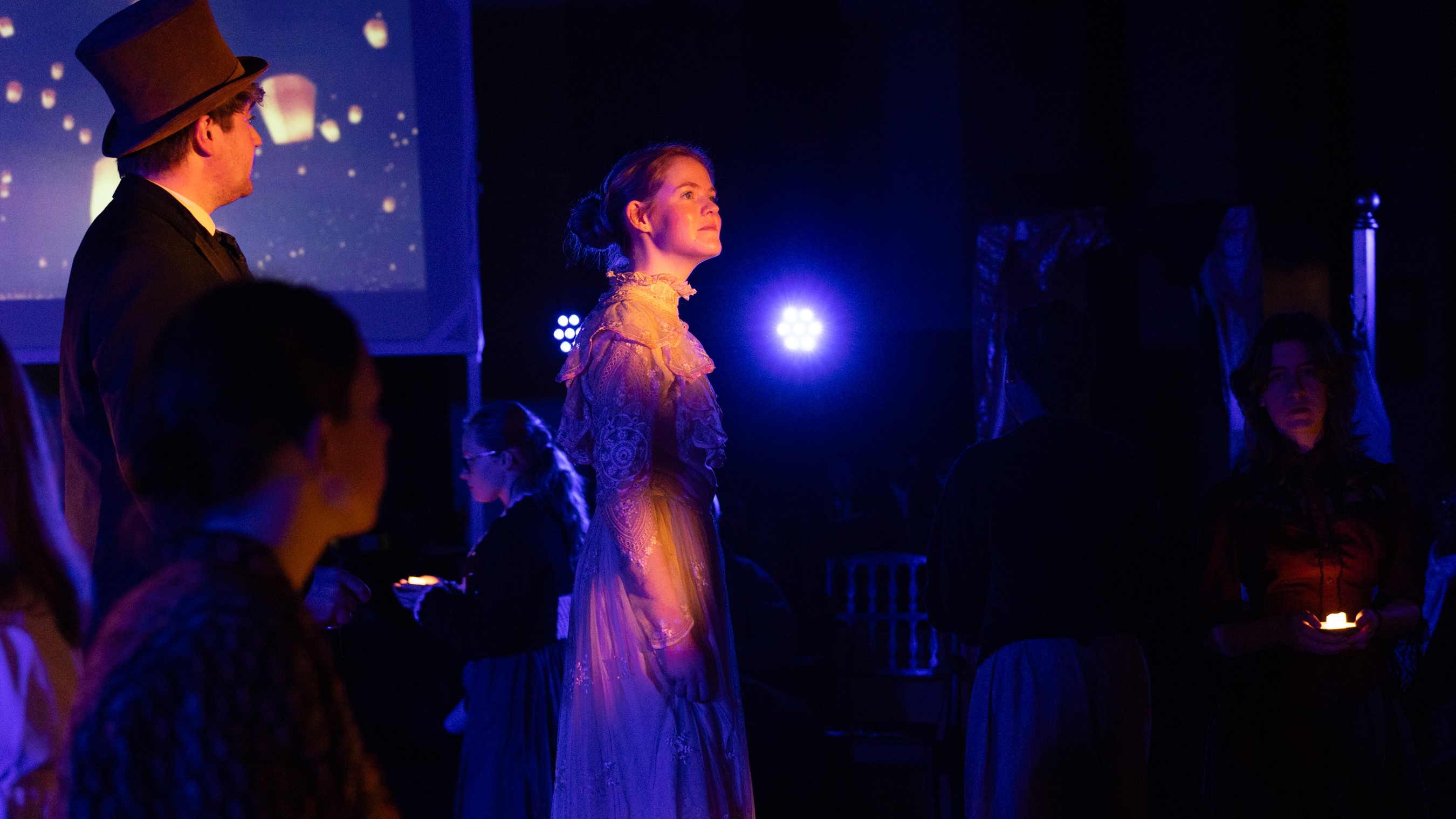 Around the World in 80 Days: a female Fogg stands on a darkened, blue-tinted stage with an image of floating paper lanterns in the background, lit by spots of golden light, looking pensive