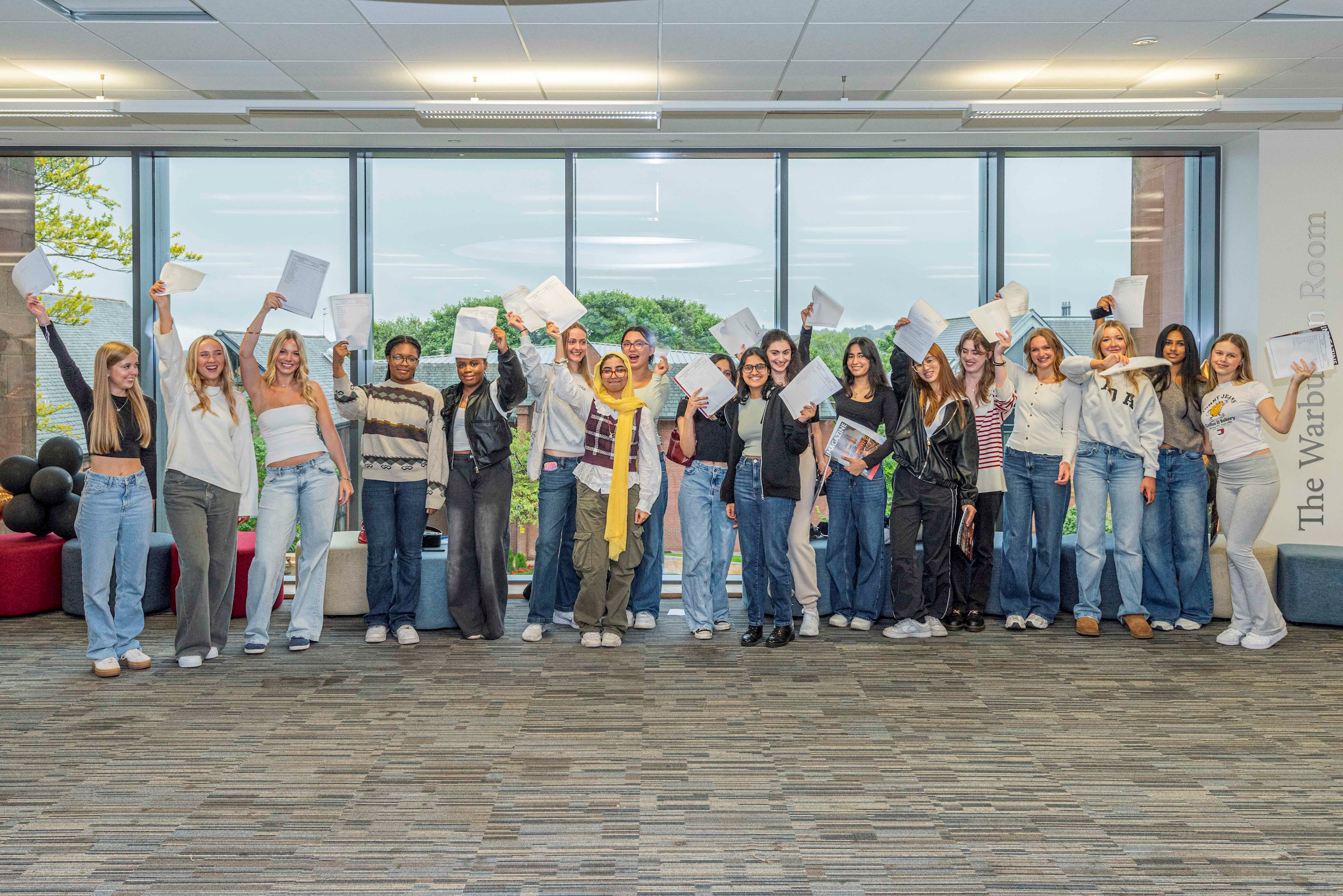A diverse group of Bolton School Girls' Division Year 11 students smiling and waving their results in the air in the common room area in the first floor of the Riley Sixth Form Centre