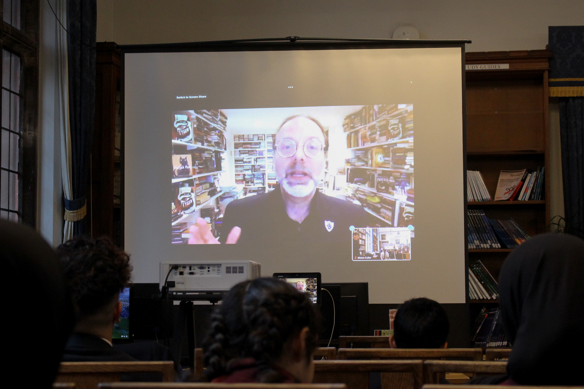 In the Boys' Division Library, a large screen has been set up in front of a group of Boys' and Girls' Division pupils, on which SF Said appears in his office with copies of his book 'Tyger' on the shelves around him