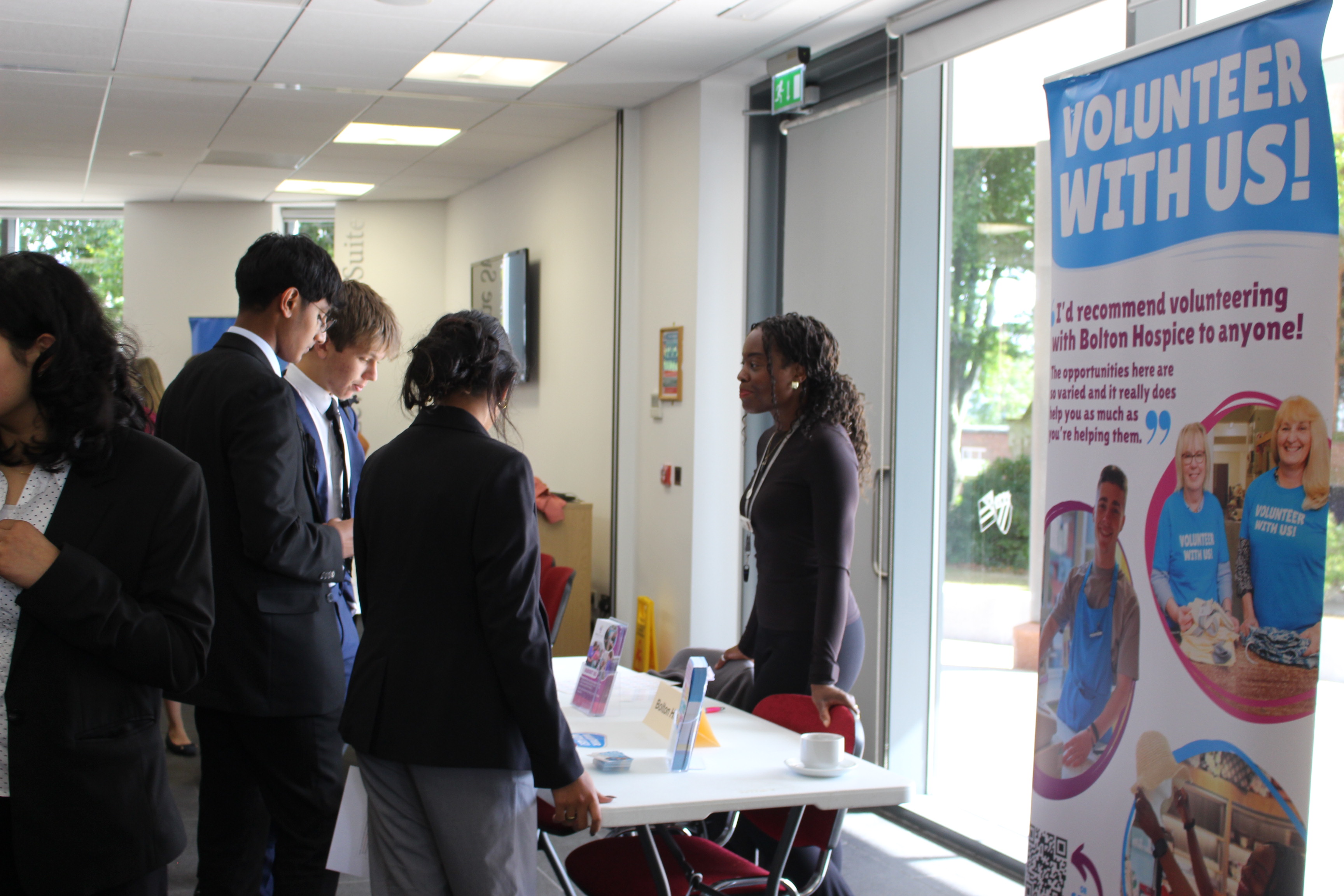 A group of Bolton School students talking to a volunteer next to a "Volunteer with us!" sign