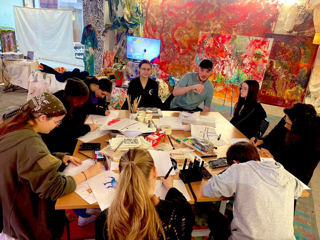 A group of teenagers clustered around a busy table, talking while making artwork, with large-scale dramatic red paintings filling the wall behind them
