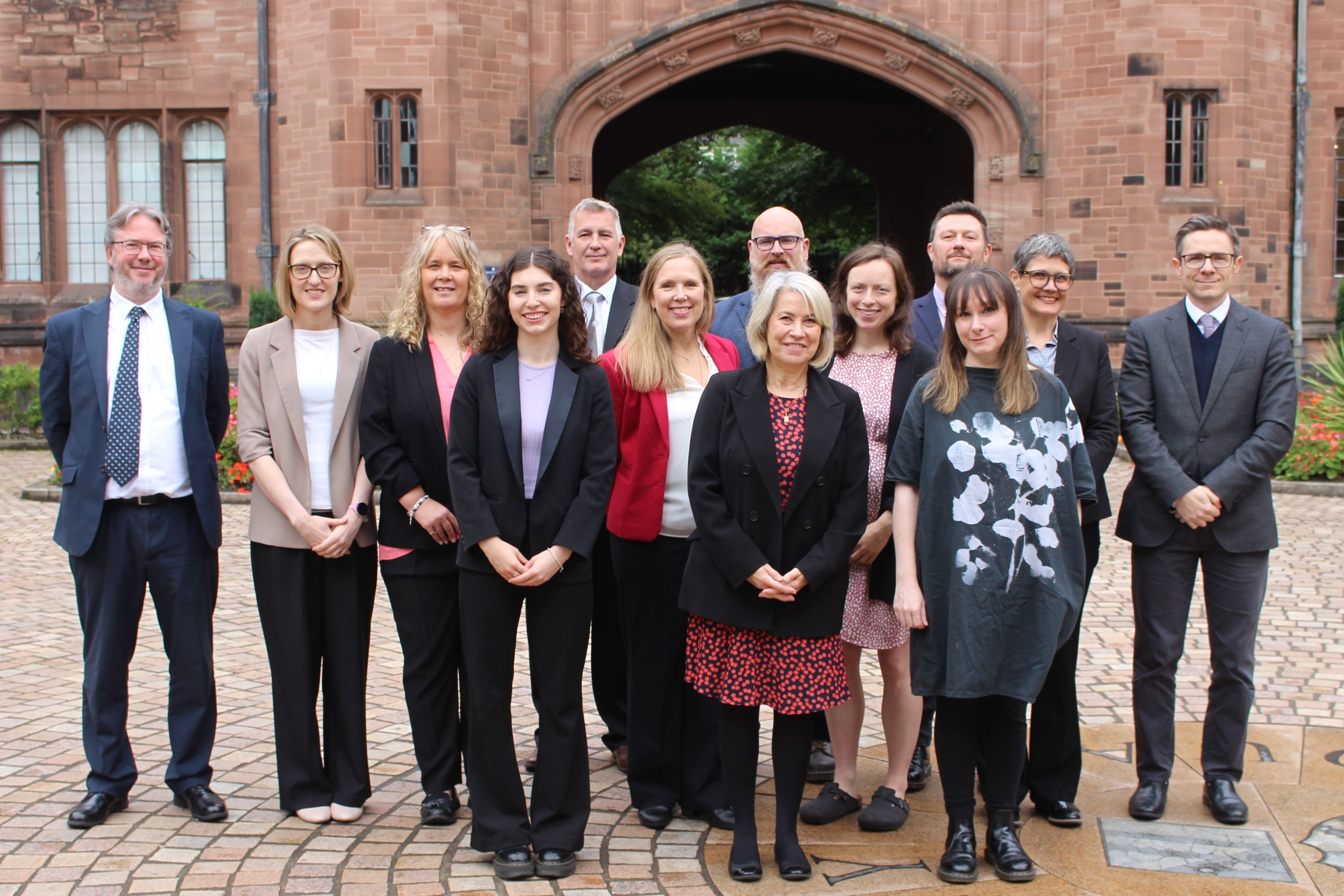 The Partnerships Team: a group of 13 smiling men and women standing outside in front of the iconic red sandstone archway that leads out of Bolton School's central Riley Quad