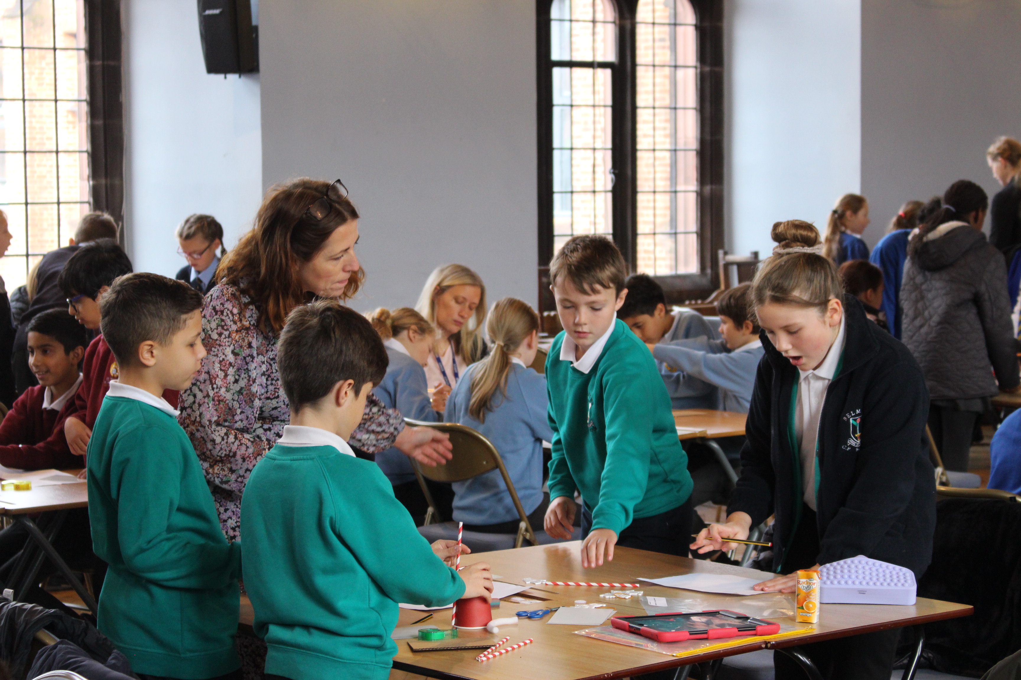 A group of primary pupils in teal jumpers taking part in an activity in a large, bright Hall with more groups around and behind them.