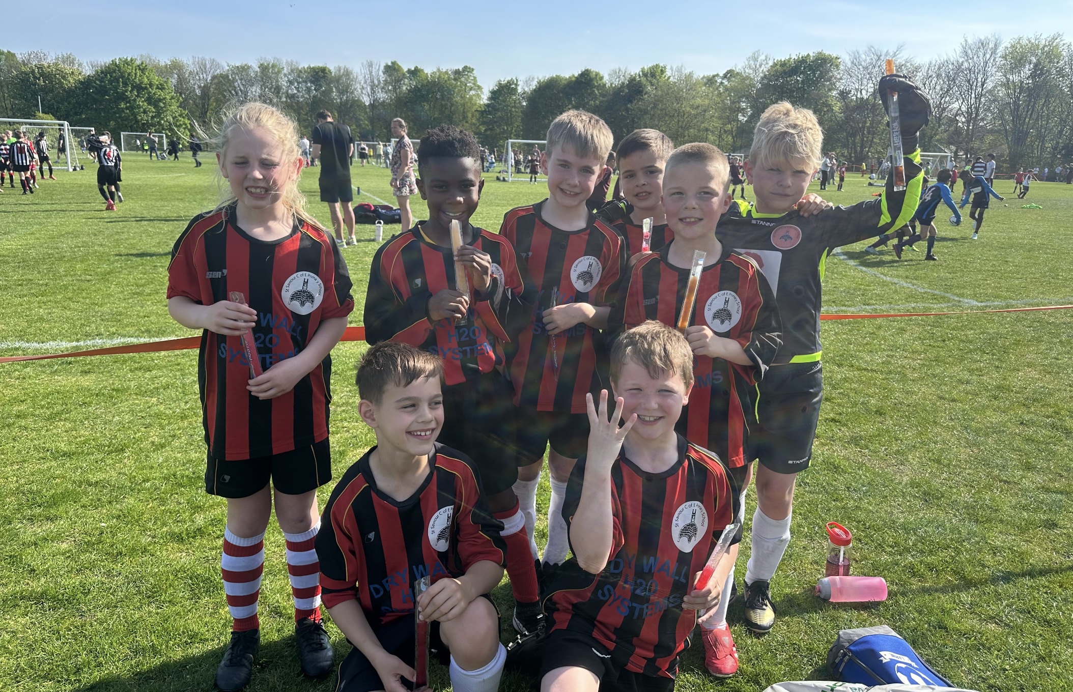 A team of boys and girls in football kit featuring red and black vertical stripes, smiling as they enjoy ice lollies while matches take place behind them